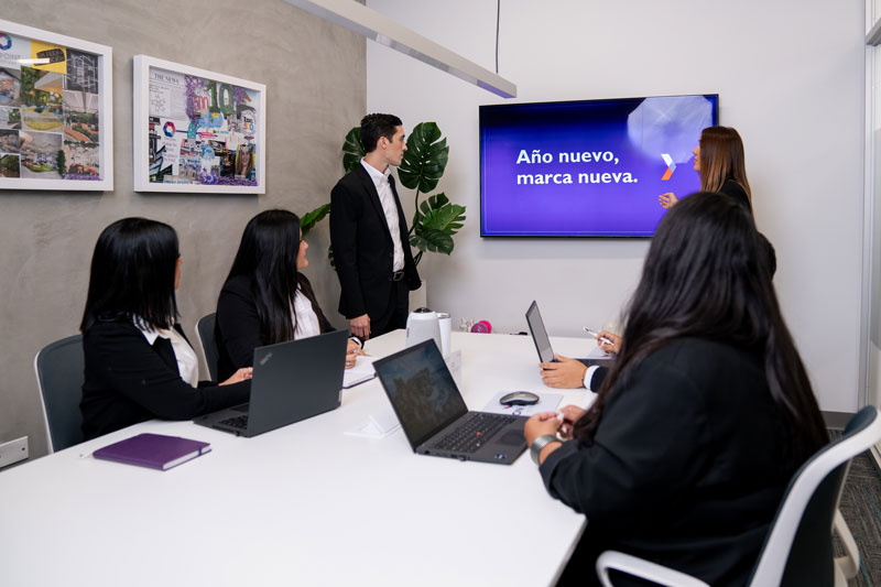 Business meeting with five professionals around a table, one standing presenting a screen displaying 'Año nuevo, marca nueva.'