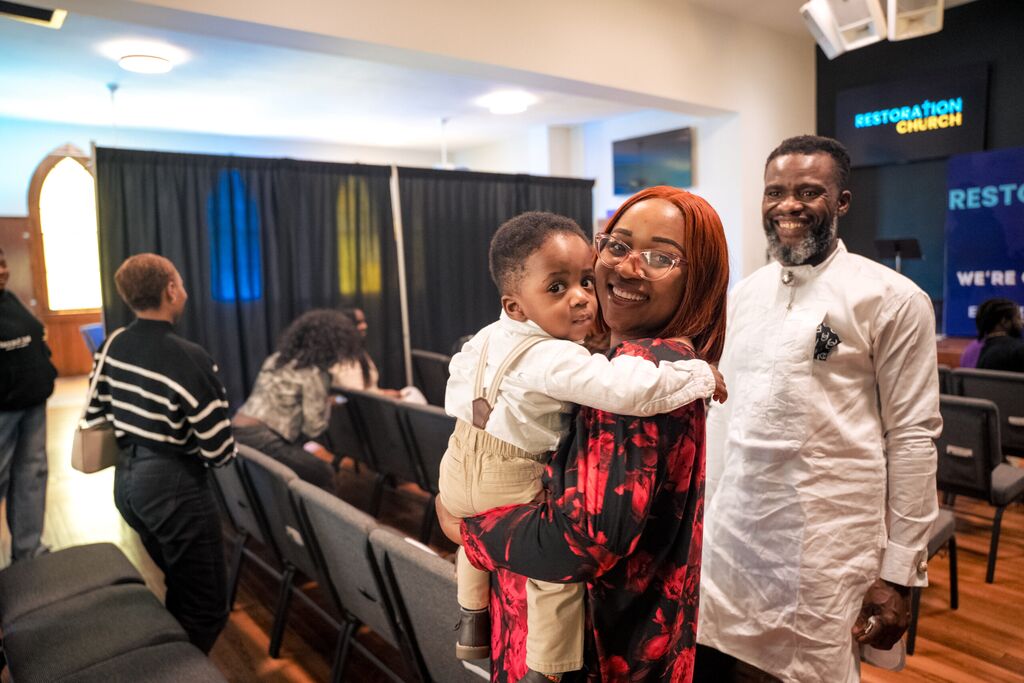 Smiling woman holding a toddler inside a church, with a man in white standing nearby and other people seated in the background.