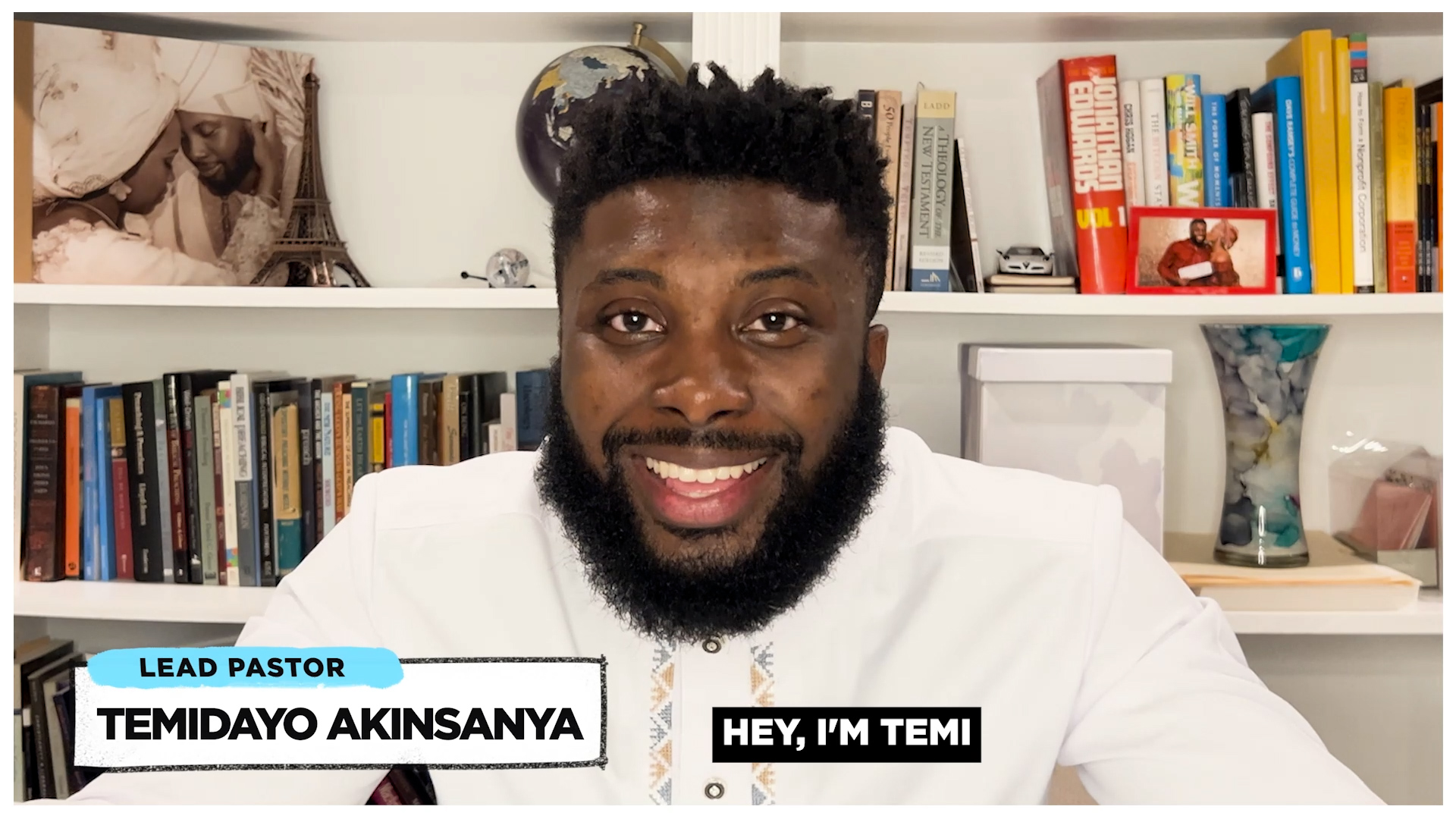 Smiling man with beard wearing a white shirt sits in front of a bookshelf full of books and a photo of a couple.