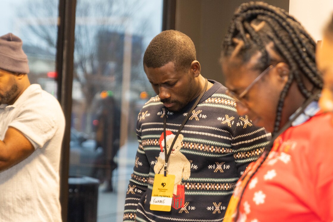 Three people standing closely indoors with heads bowed, one wearing a festive sweater with a teddy bear and a name tag reading 'Kwame'.
