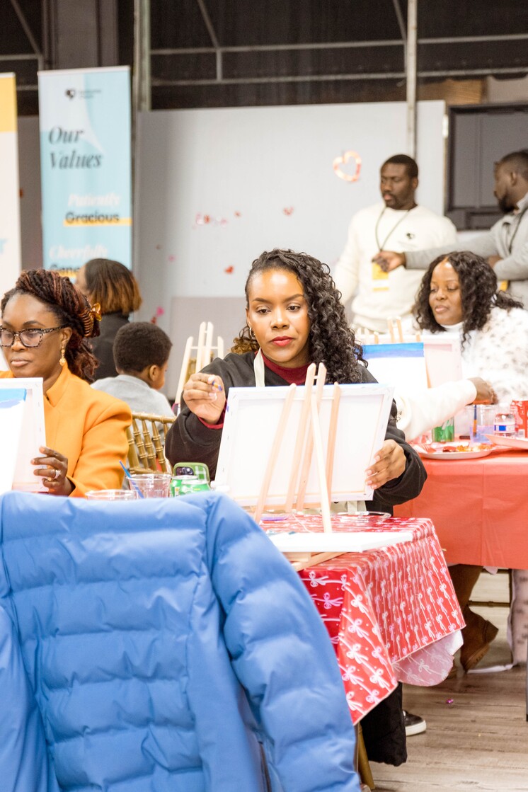 People seated at tables covered with red tablecloths, painting on canvases during a group art activity.