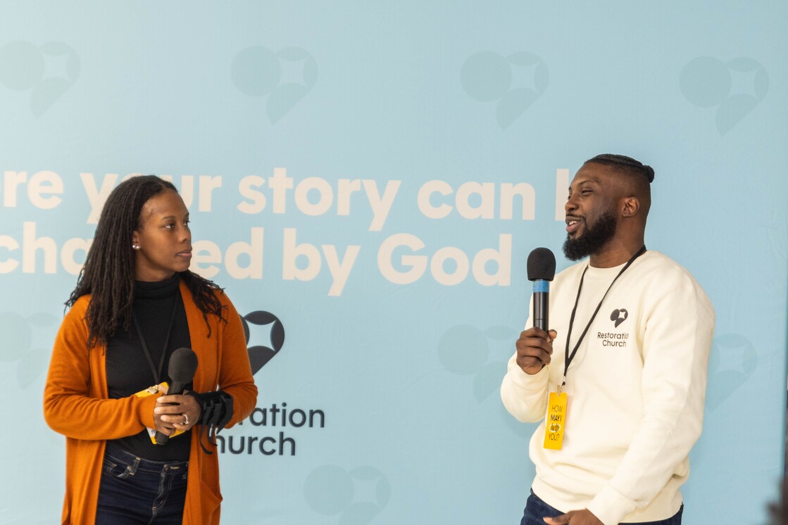 Man and woman holding microphones speaking in front of a blue backdrop with text related to Restorations Church.