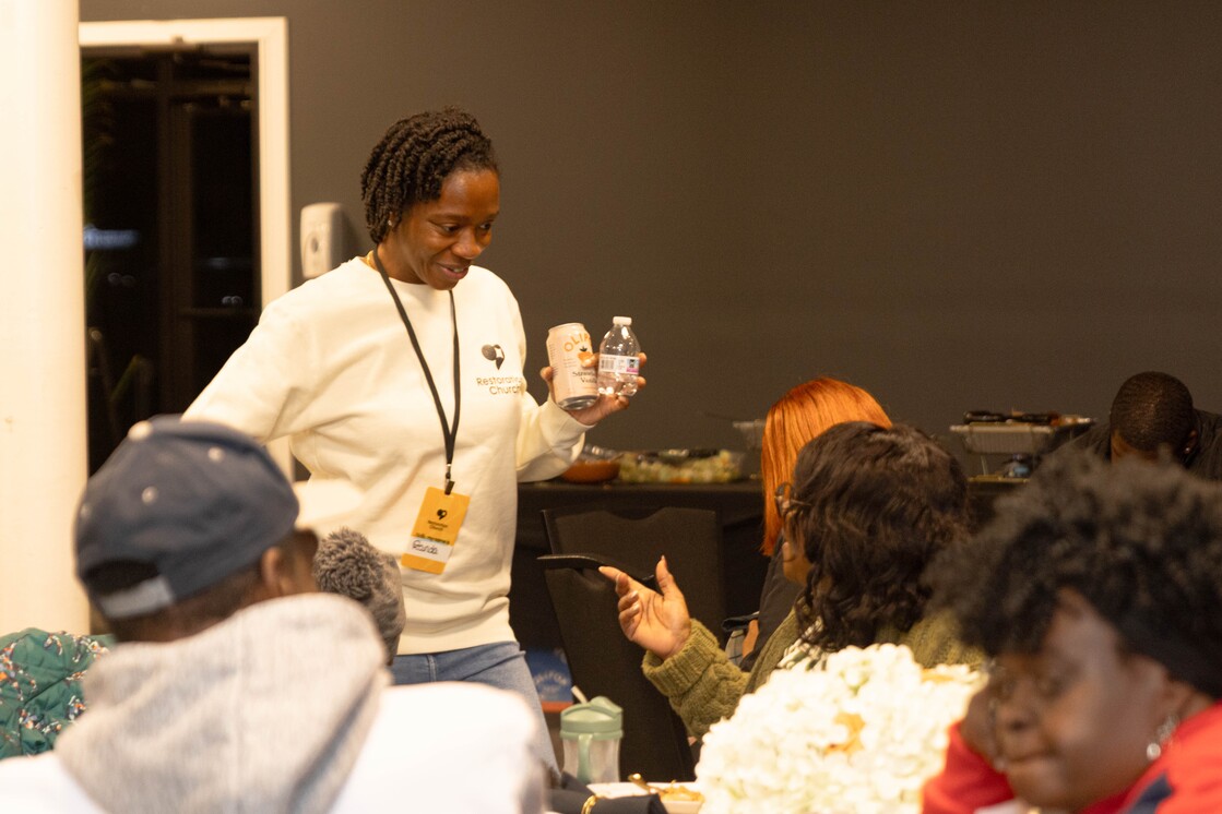 A woman holding a canned drink and a water bottle while engaging with a seated group in a social setting.