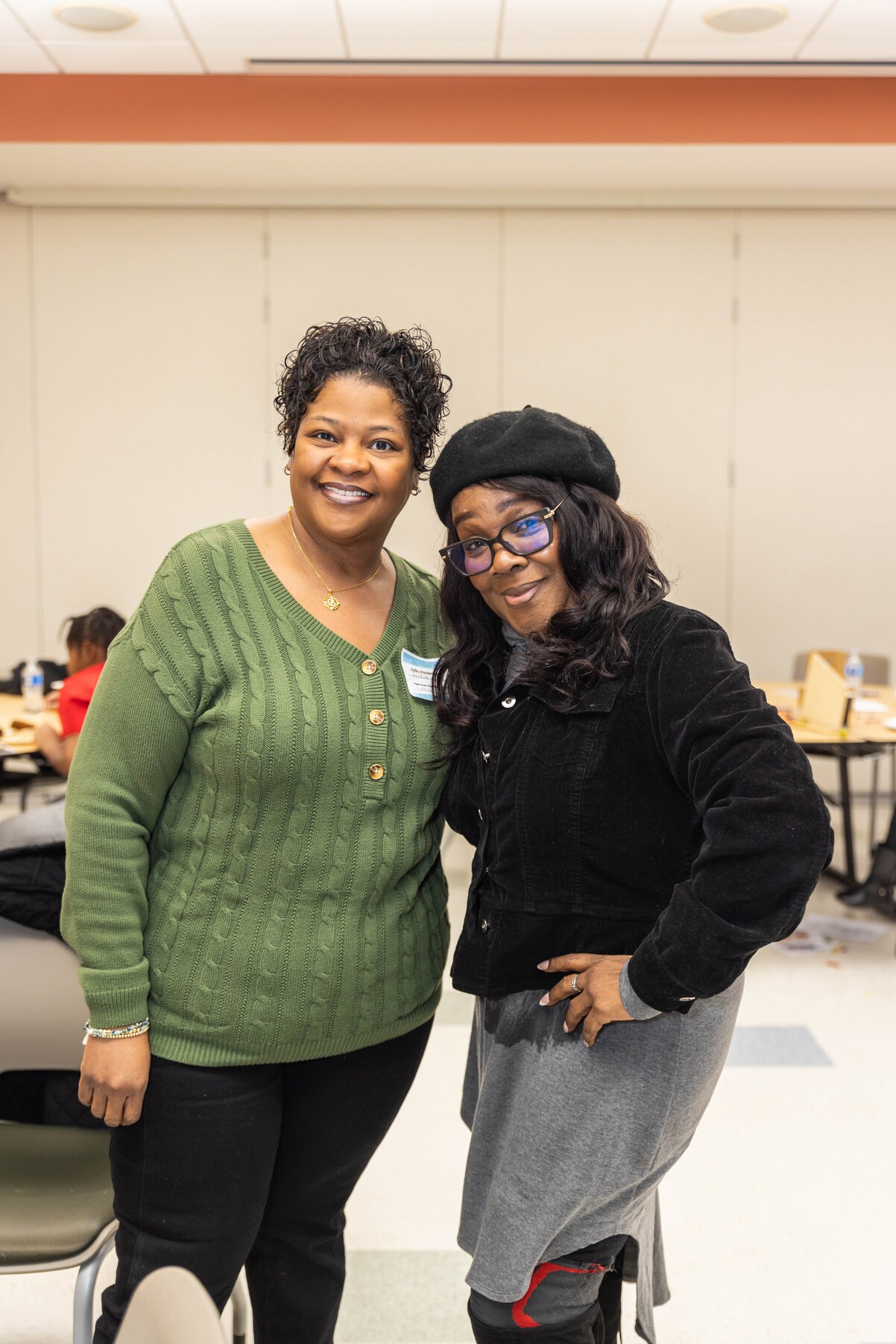 Two smiling women posing together indoors; one in a green sweater and the other in a black jacket and beret.