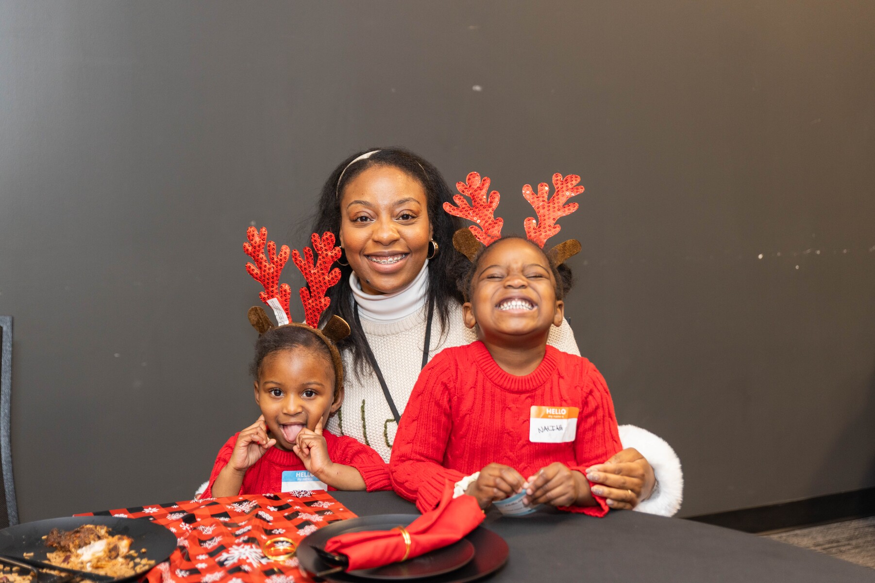 Smiling woman with two children wearing red sweaters and red reindeer antler headbands at a holiday table.
