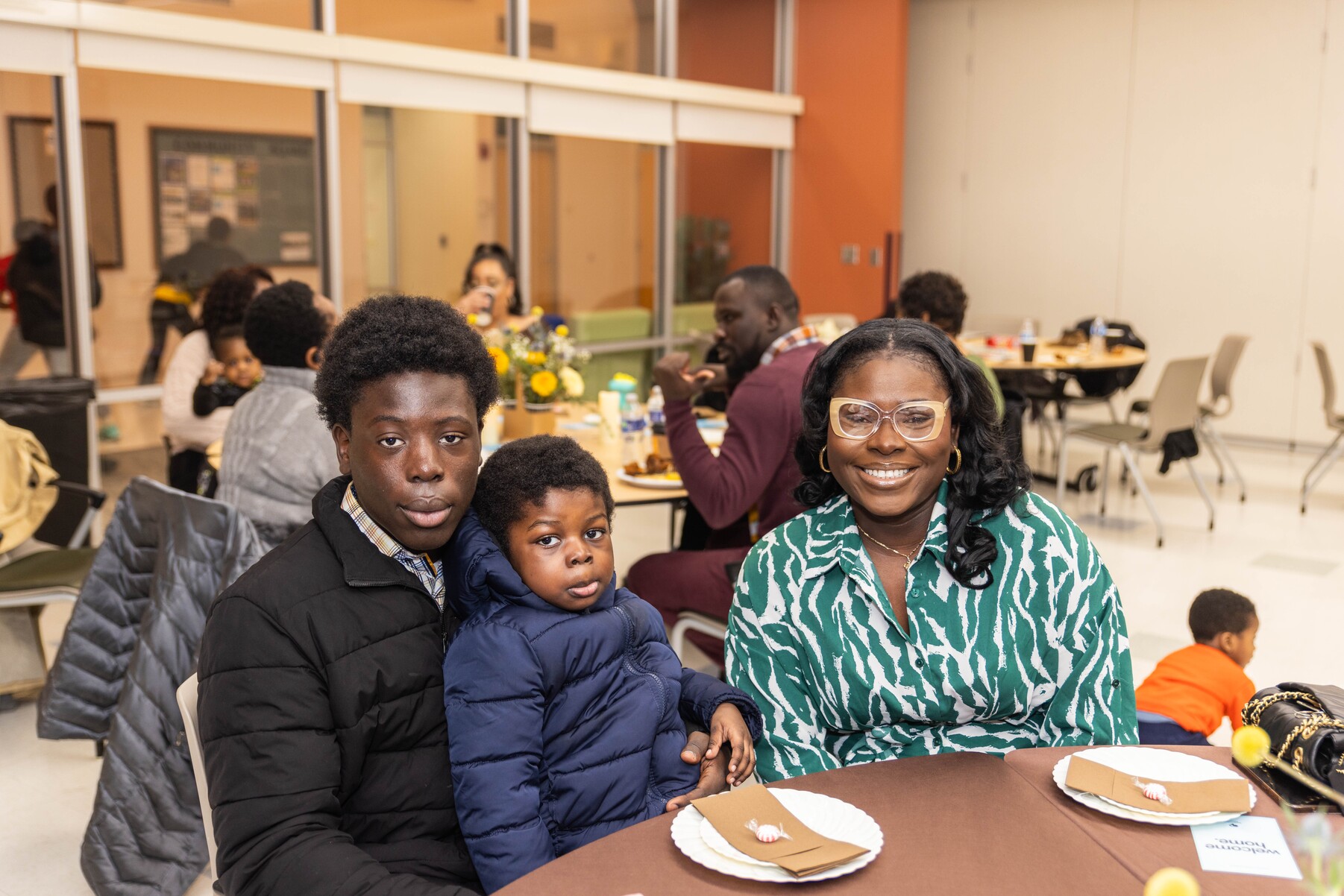 A smiling woman wearing glasses and a green patterned shirt sits at a table with two boys, one holding the younger on his lap, in a well-lit room with other people dining.