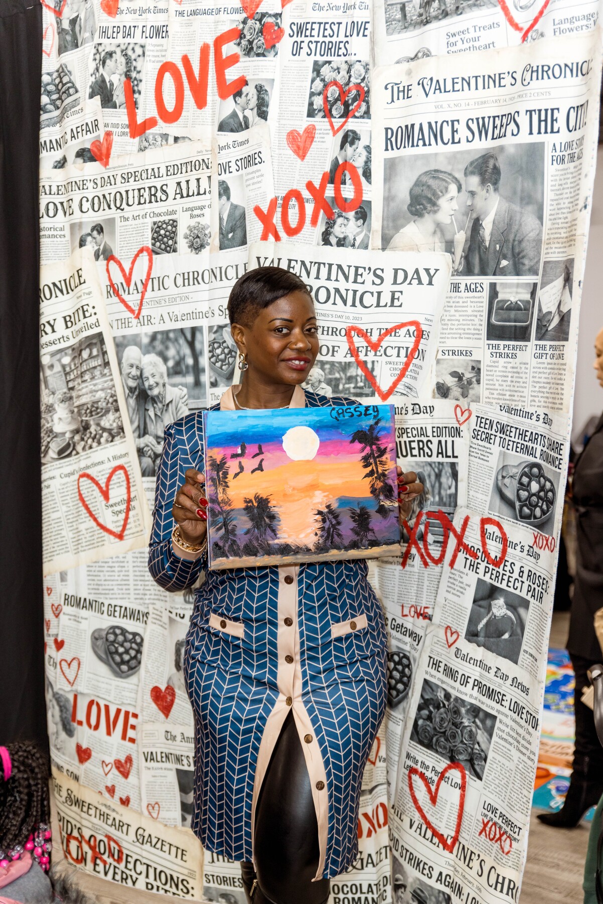 Woman in blue patterned dress holding a colorful painting of a sunset with palm trees and birds, standing in front of a Valentine's Day themed backdrop with red hearts and newspaper-style text.