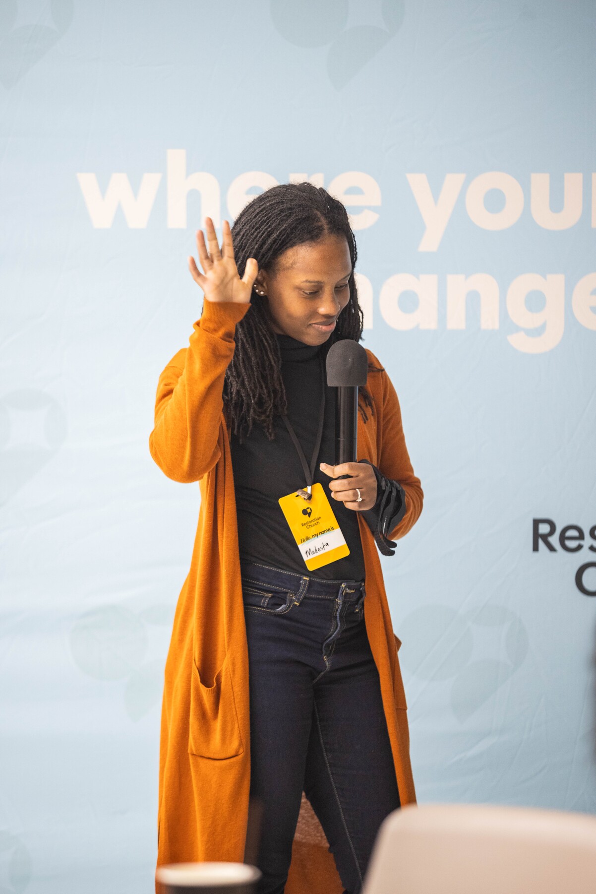 Woman in orange cardigan holding a microphone and raising her hand, standing in front of a blue backdrop with partial text.