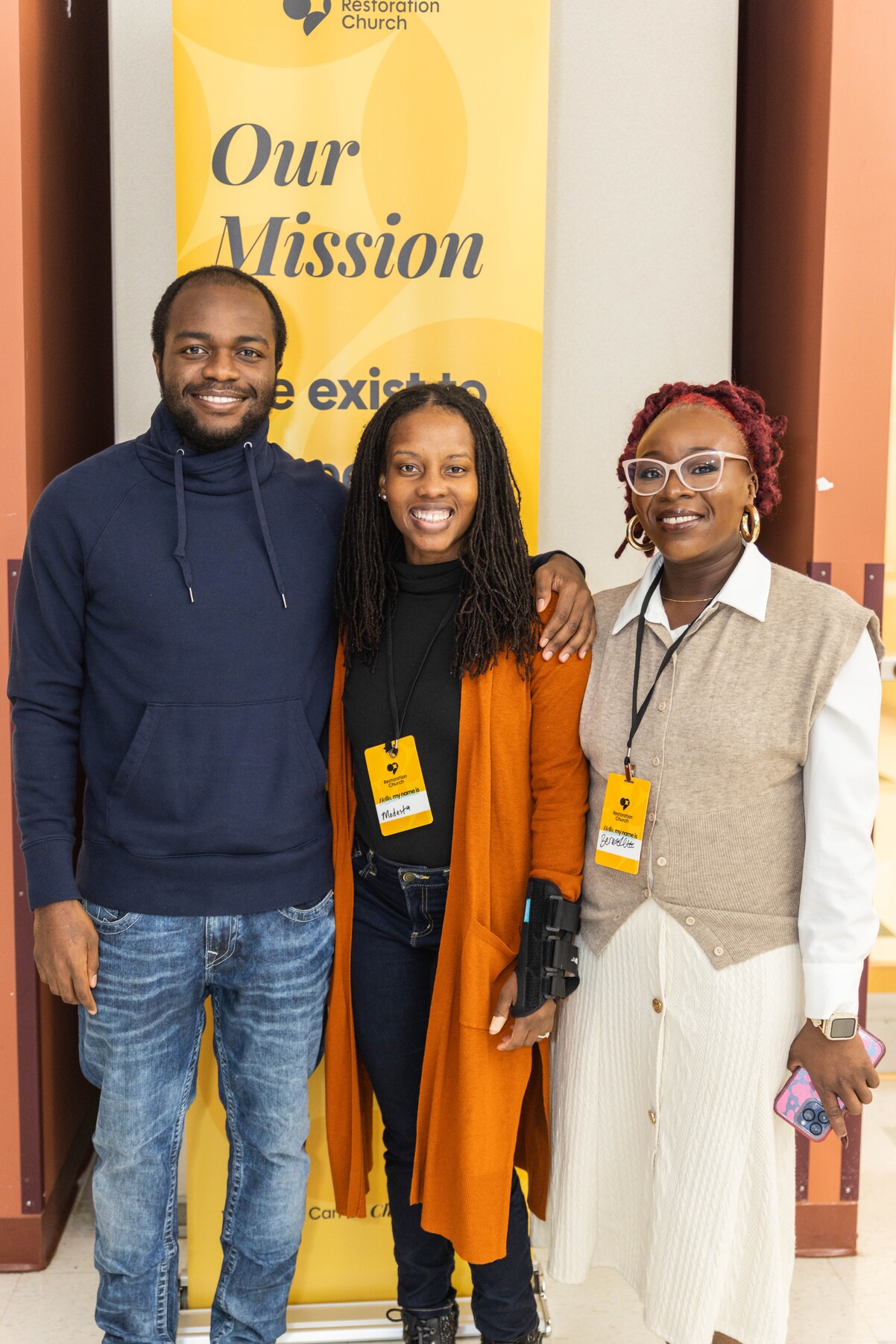 Three smiling people standing with arms around each other in front of a yellow Restoration Church mission banner.