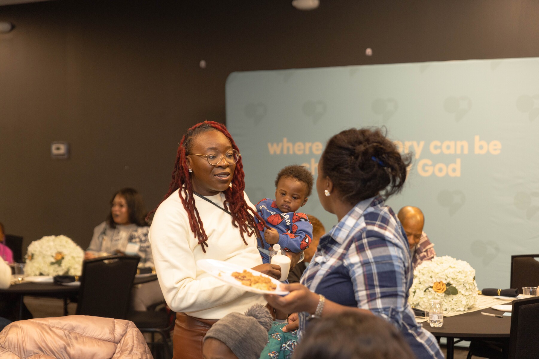 A woman with red dreadlocks holding a baby in a Spider-Man outfit talks to another woman in a plaid shirt at an indoor gathering.