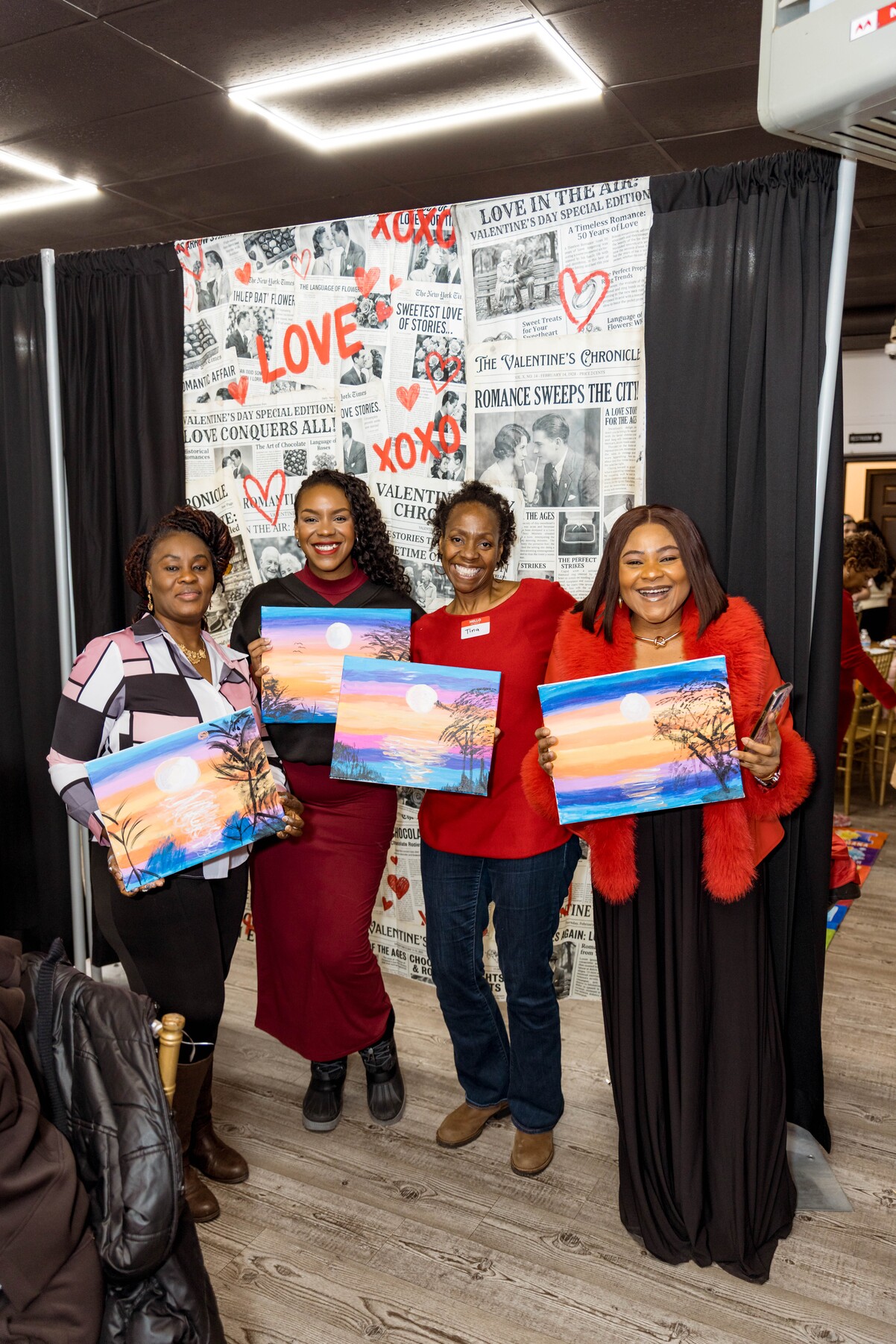 Four smiling women holding sunset landscape paintings in front of a Valentine’s Day themed backdrop.