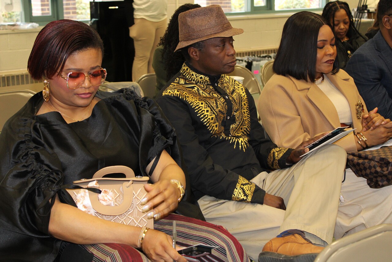 Three people sitting indoors on chairs, with one woman wearing sunglasses and holding a beige purse, a man wearing a brown hat and patterned black and gold shirt, and another woman in a beige coat holding a booklet.