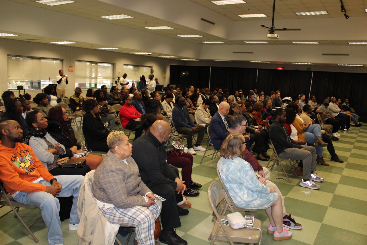 Large group of people seated in rows of chairs attentively listening to an event in a conference room with green and beige checkered floor.