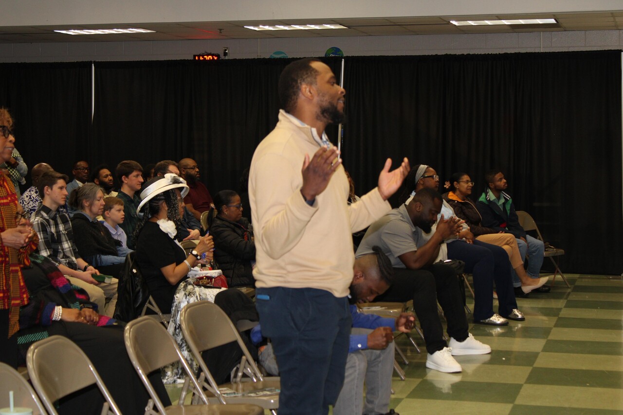 Man standing with raised hands among a seated diverse group in a room with green checkered floor and black curtains.