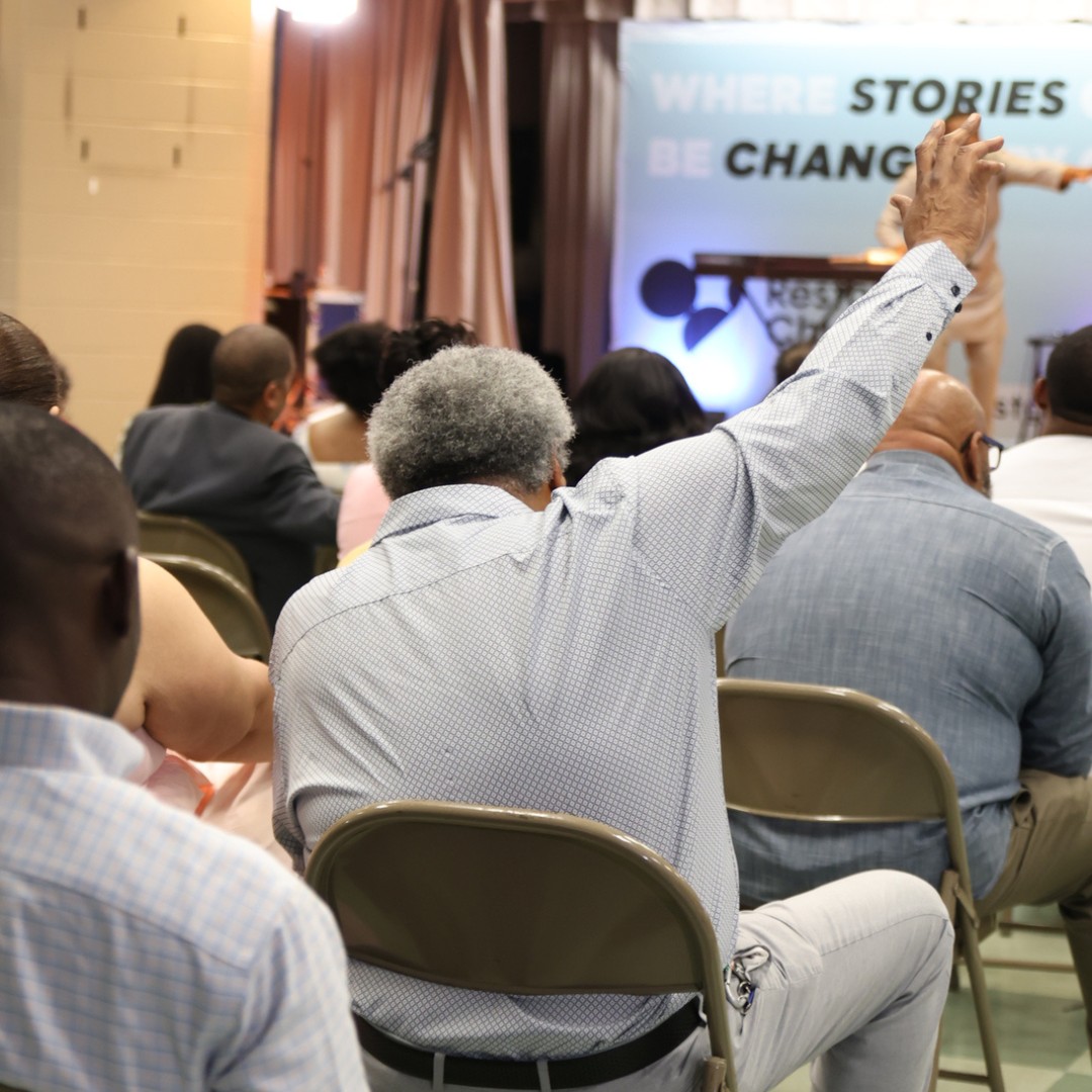 Man with gray hair raising his hand while sitting in an audience facing a speaker on stage with a backdrop that reads 'WHERE STORIES BE CHANGE'.