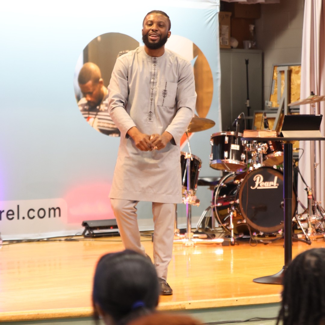 Man in traditional attire speaking on a stage with drums and a microphone stand behind him.