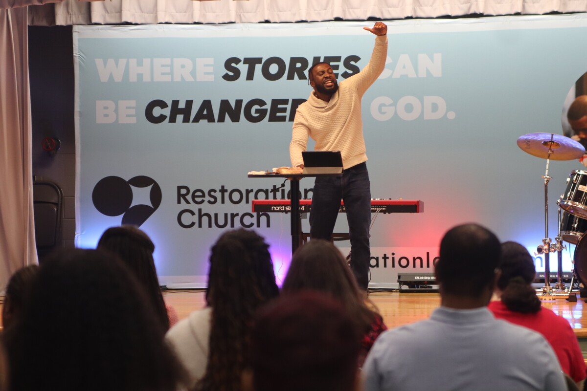 Speaker passionately raises hand addressing seated audience at Restoration Church worship event.