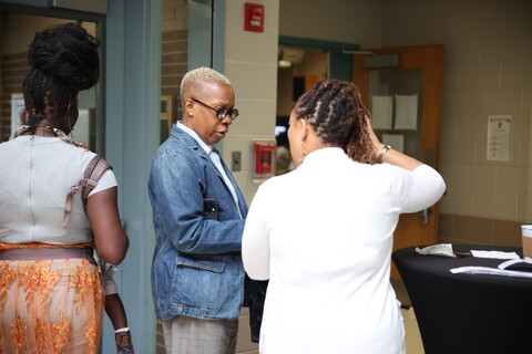 Three people engaged in conversation in a hallway, with one person holding a pen and paper.
