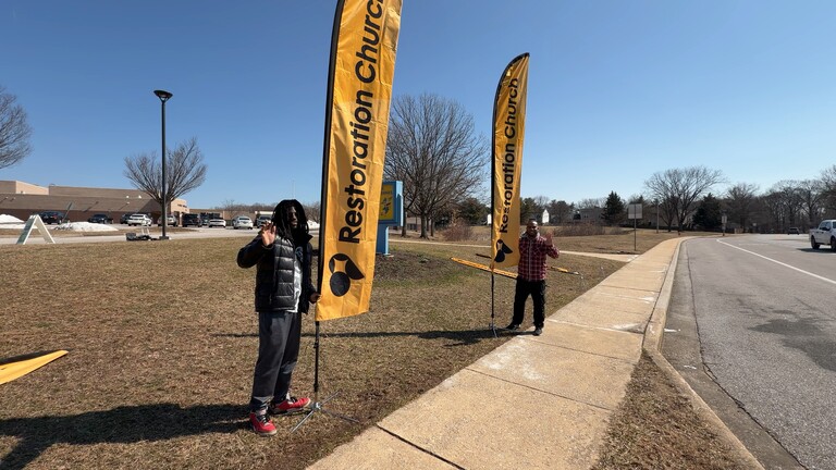 Two men waving near yellow Restoration Church flags on grass by a sidewalk and street on a clear day.
