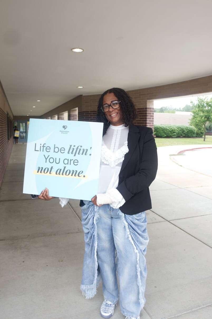Woman holding sign saying 'Life be lifin': You are not alone' standing under a covered walkway.