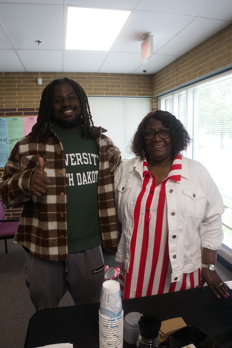 Smiling man in plaid jacket and woman in striped dress stand behind table with cups indoors.