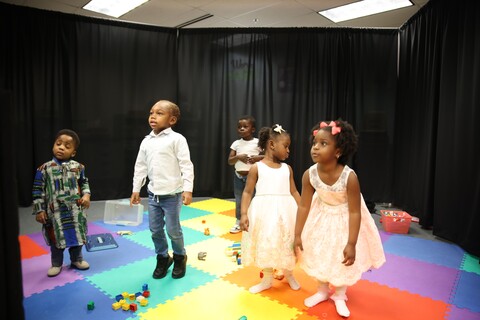 Five children playing on colorful foam mats with toys in a room with black curtains.