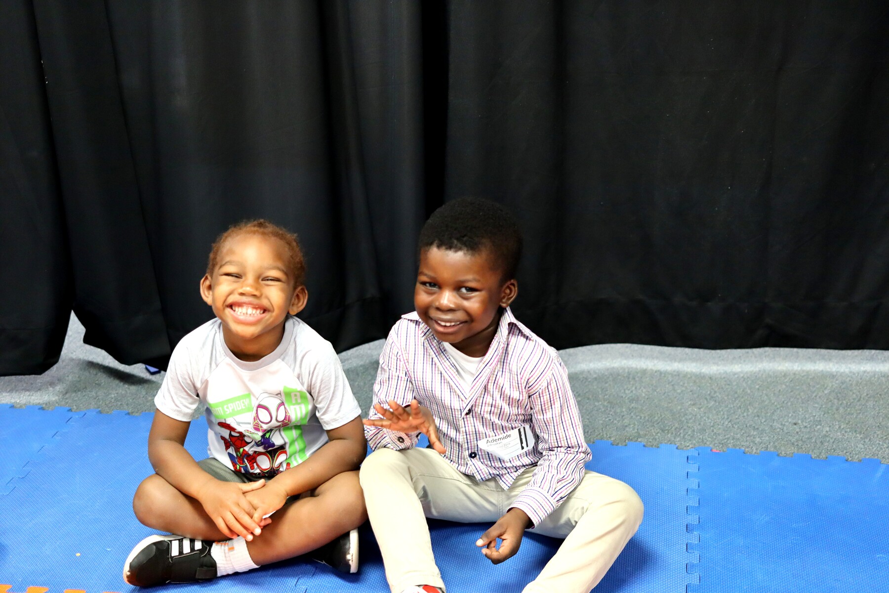 Two young boys sitting on blue mats; one smiles broadly, the other waves hand.