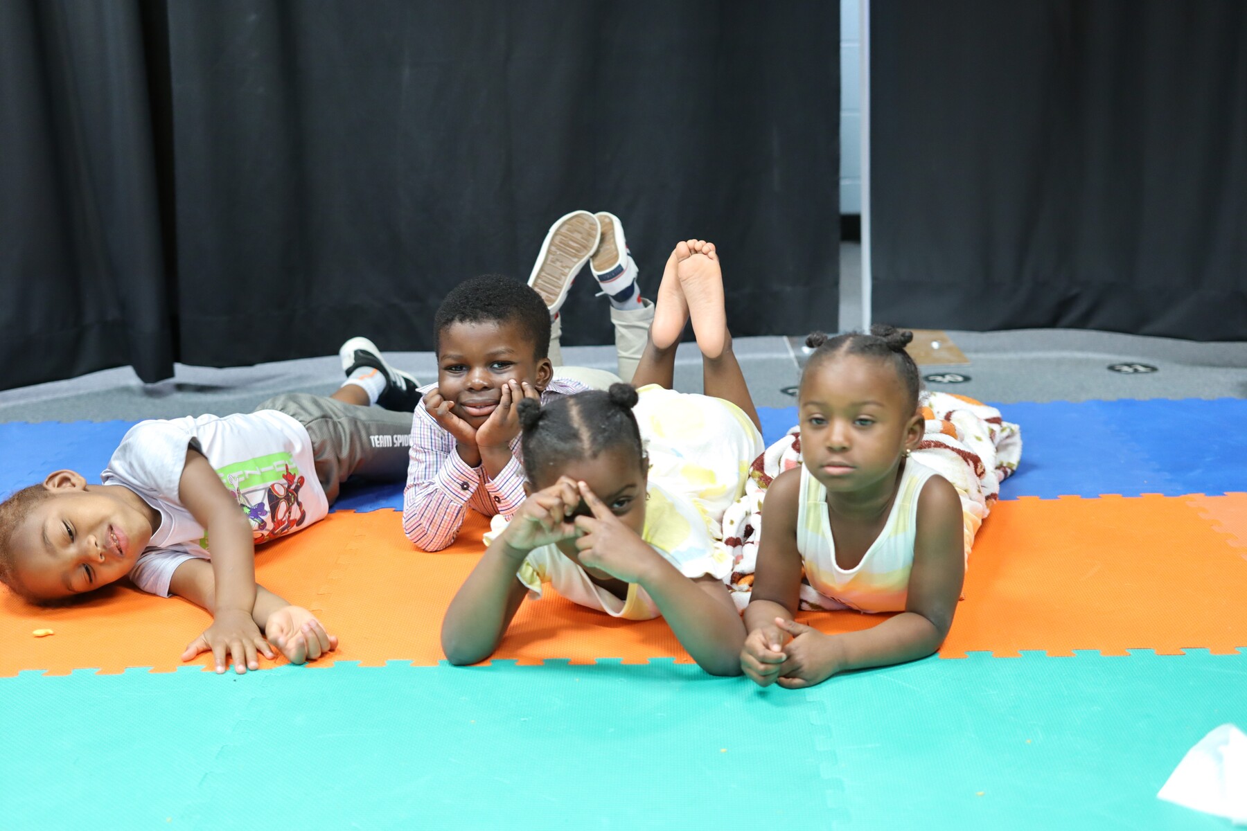 Four children lying on colorful floor mats, two with hands on their faces, one making a triangle with fingers.