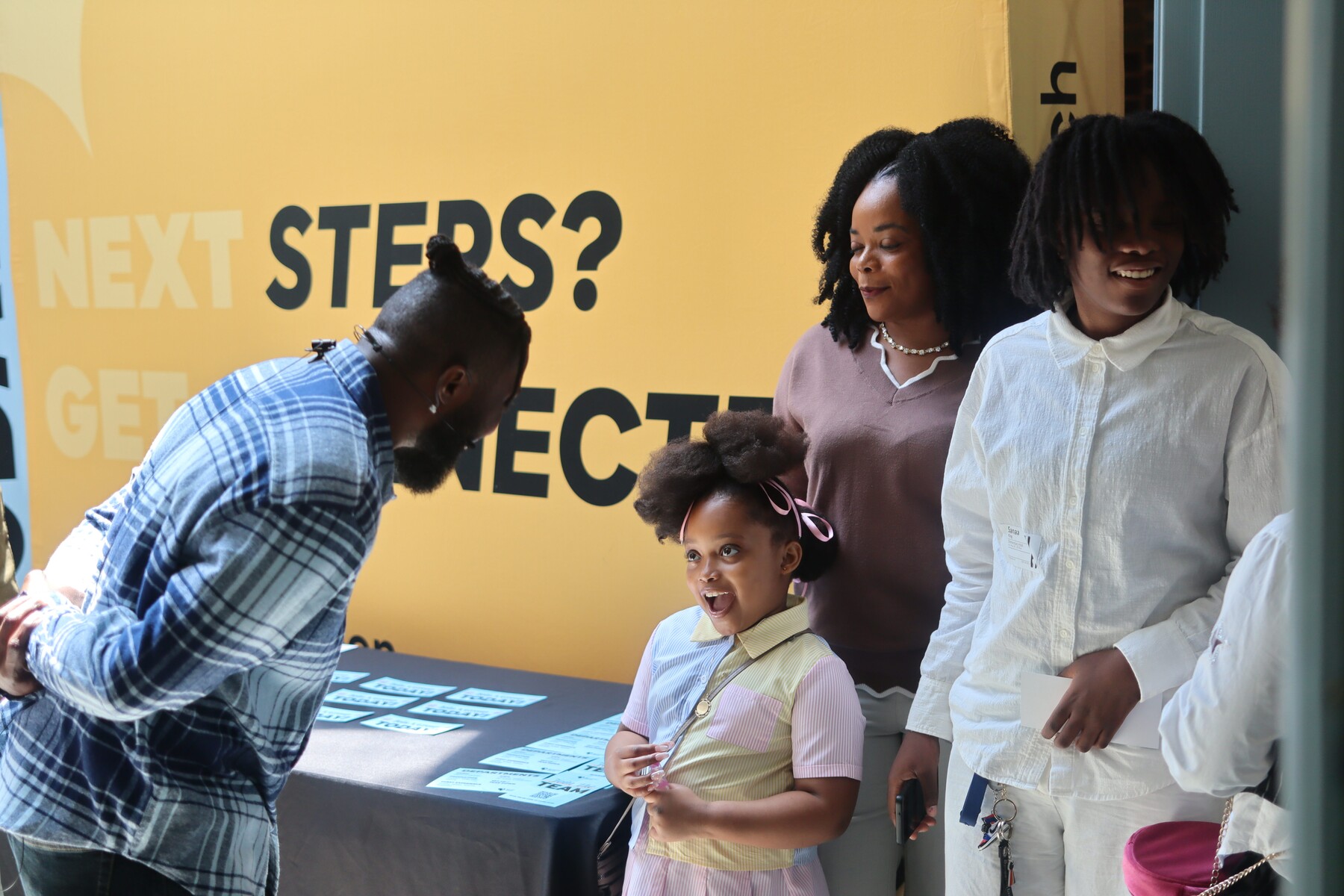 Man in plaid talks to smiling young girl, woman, and boy in front of a yellow sign reading 'Next Steps?'