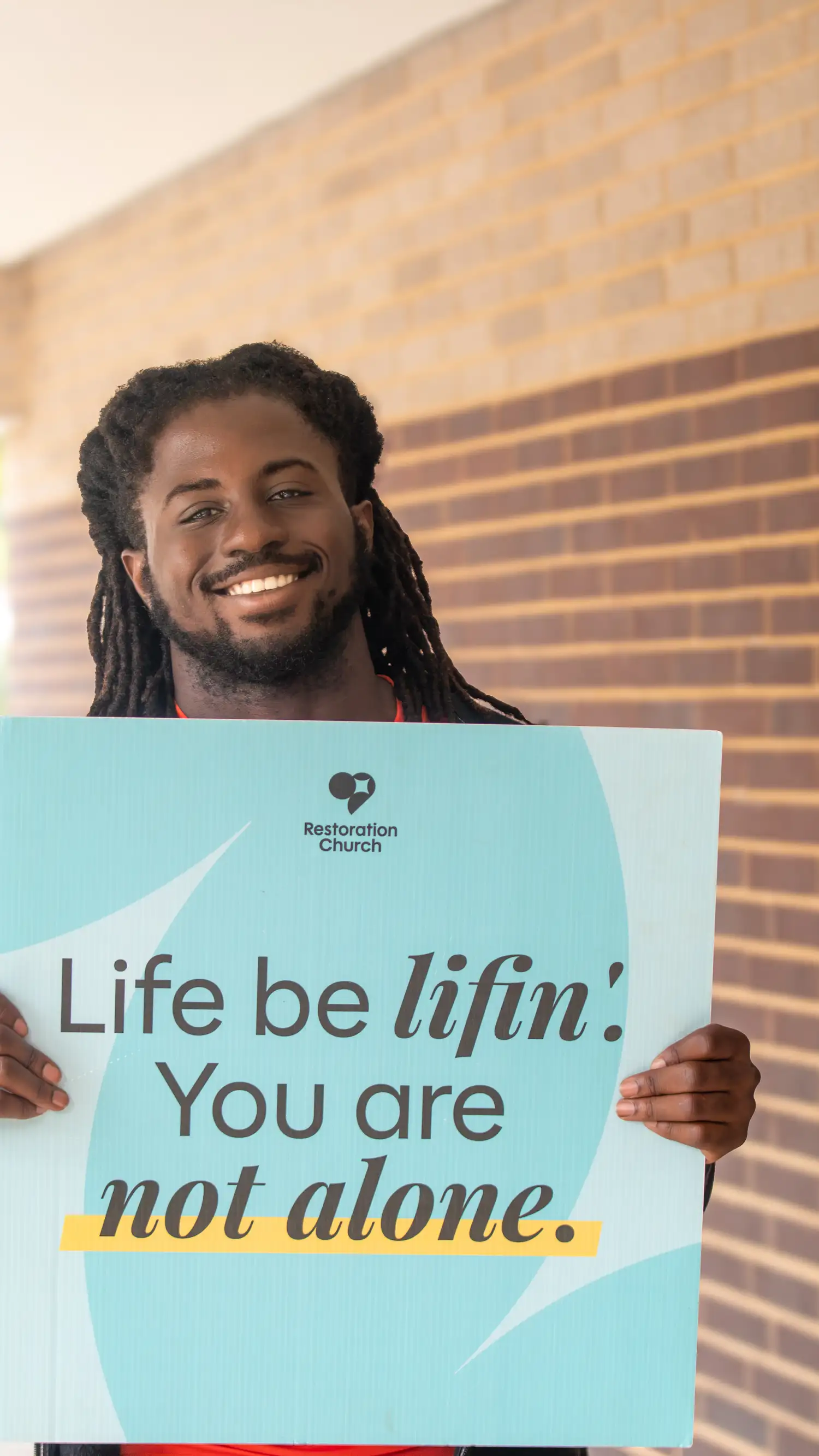 Smiling man holding a Restoration Church sign that reads "Life be lifin'! You are not alone."