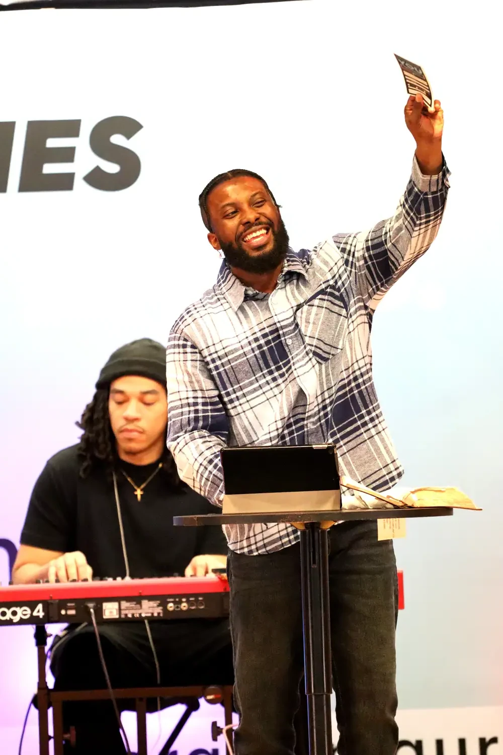 Smiling speaker holding up a card at the podium while a keyboardist plays at Restoration Church in Laurel, Maryland