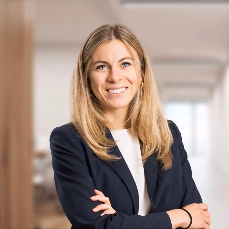 Lächelnde Frau mit langen blonden Haaren und schwarzem Blazer vor hellem Bürohintergrund.