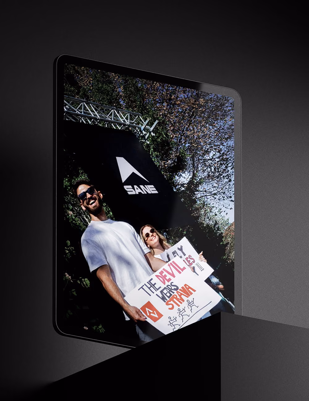 Zwei Personen mit Sonnenbrillen halten ein Schild mit dem Text ‚The devil wears Strava‘ bei einem Event.