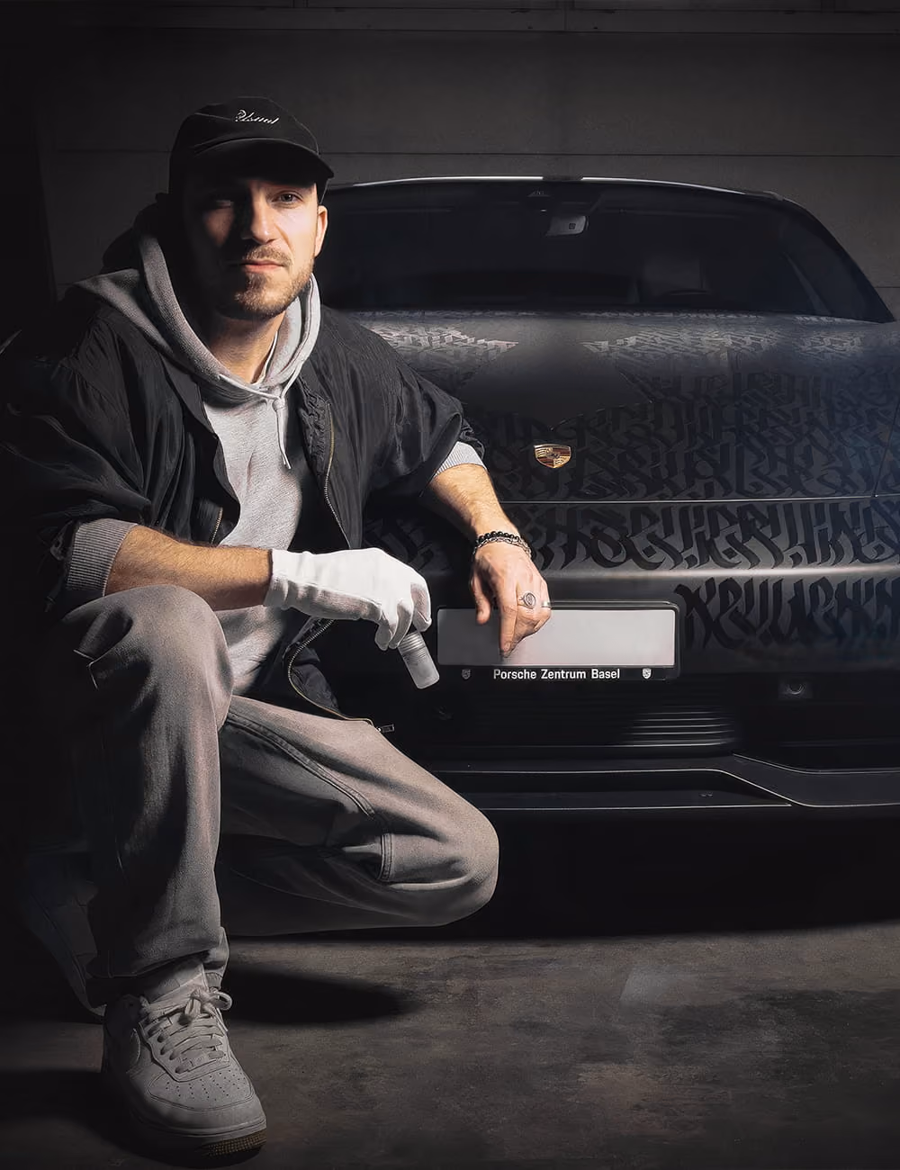 Mann in grauer Kapuzenjacke und weißem Handschuh kniet vor einem Porsche mit kalligrafischem Design.
