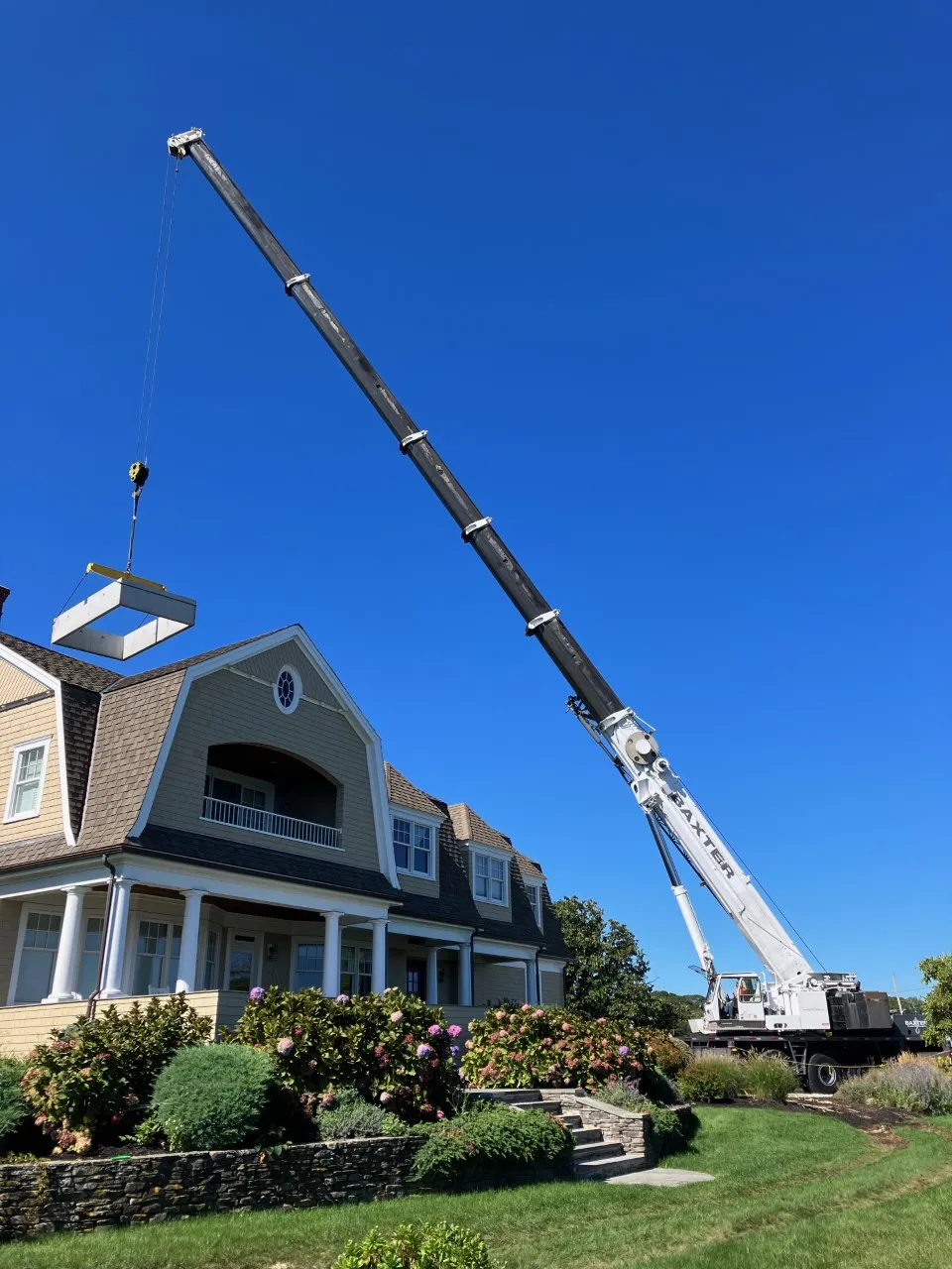 Large crane lifting a rectangular structure near a beige two-story house with a landscaped garden and clear blue sky.