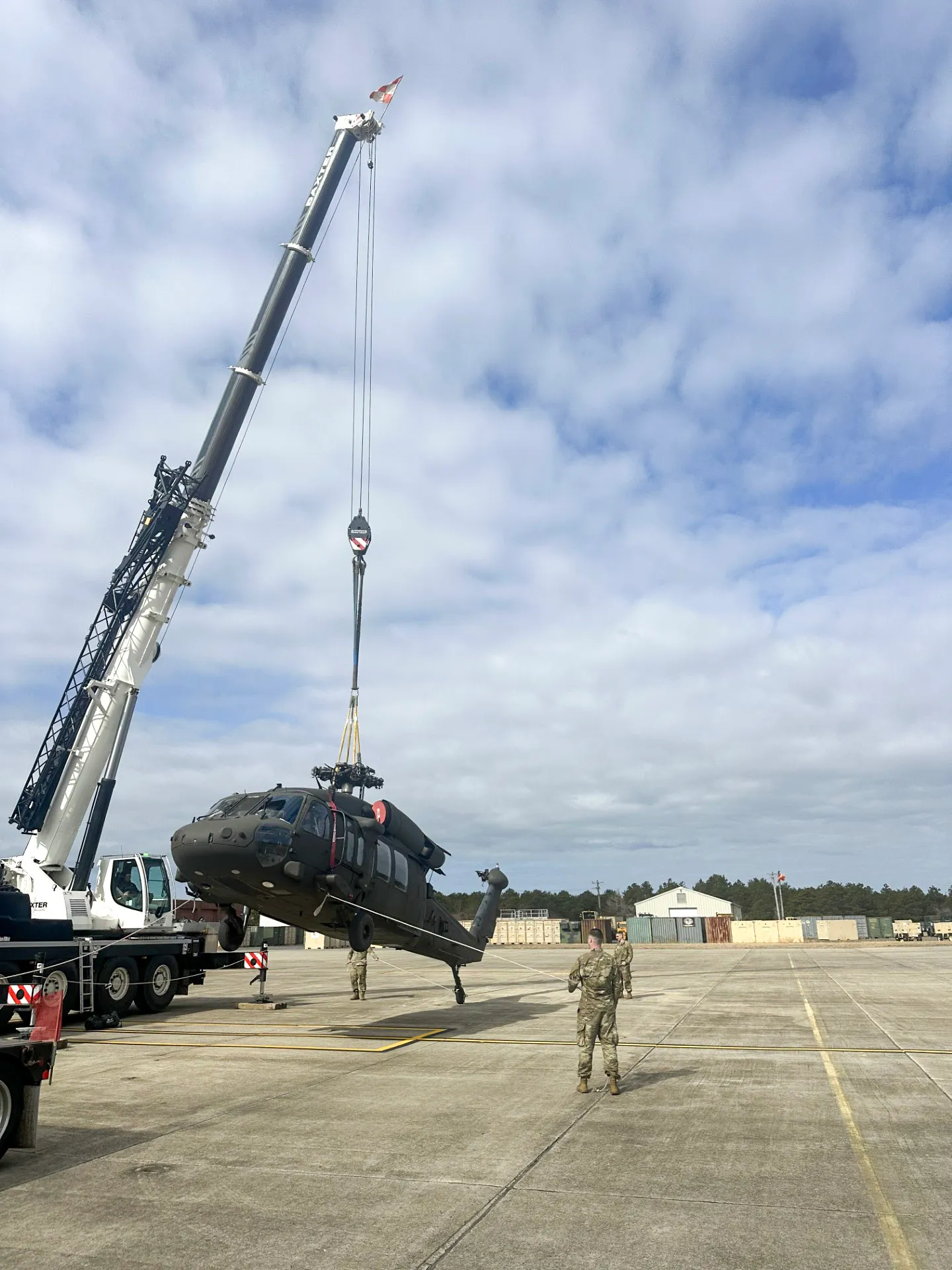 Military helicopter being lifted by a large crane on an airfield with soldiers guiding the operation.