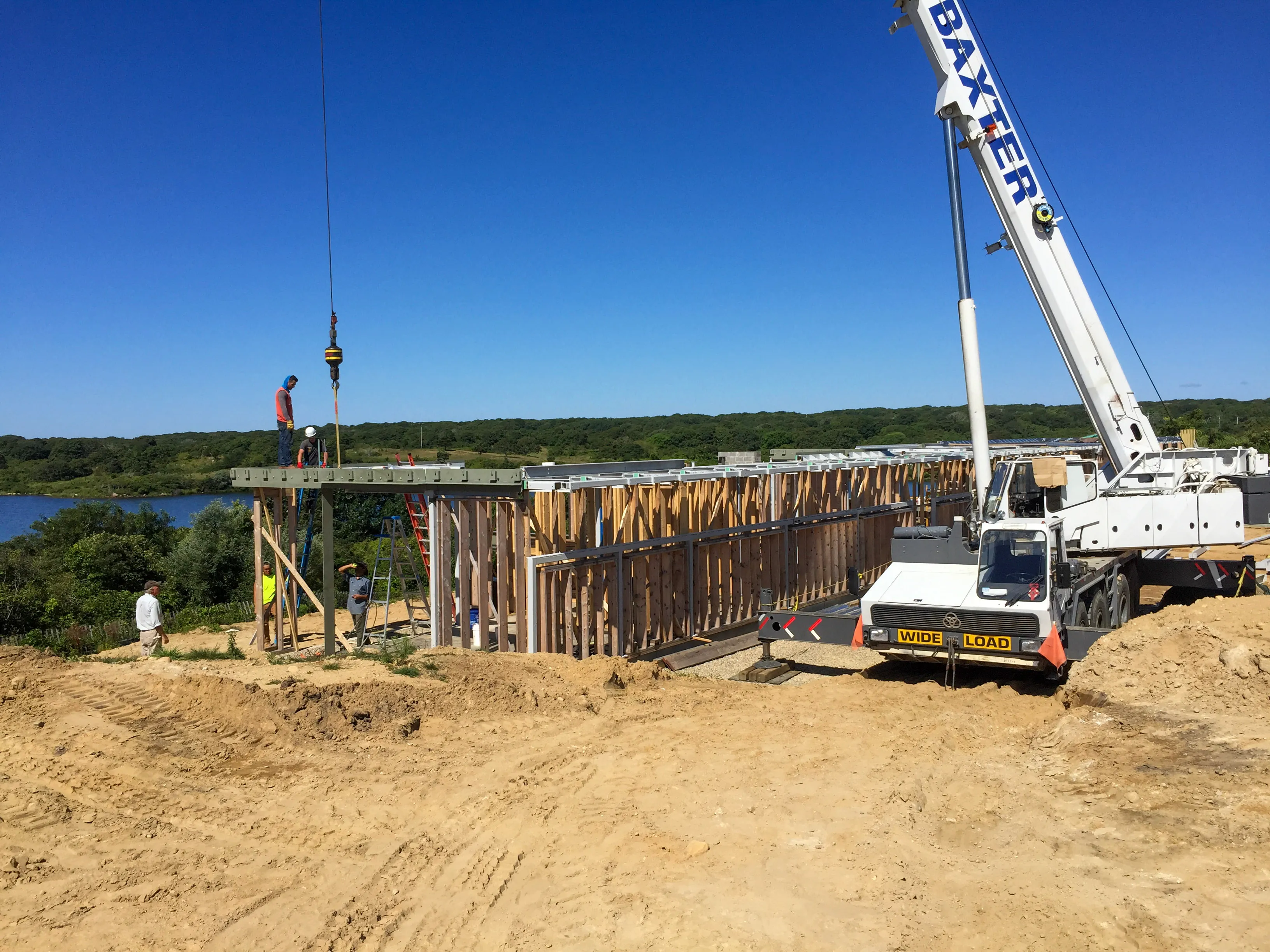 Construction site with a large white crane lifting materials onto a wooden frame structure, workers present under a clear blue sky.