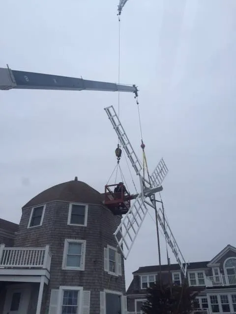 Crane lifting the sails of a traditional wooden windmill next to a shingled building on a cloudy day.