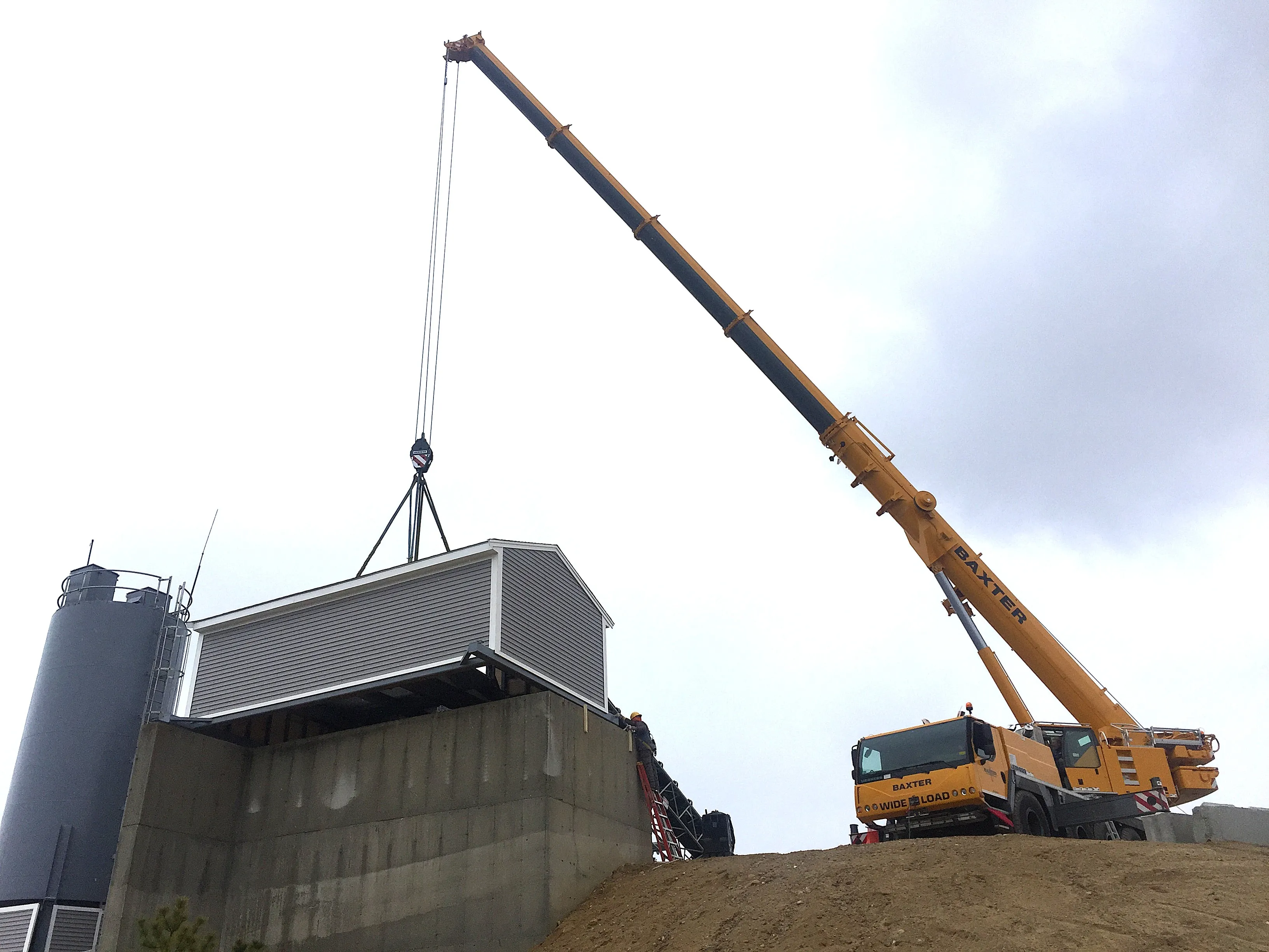 Yellow crane lifting a small gray building onto a concrete platform next to a large silo under a cloudy sky.
