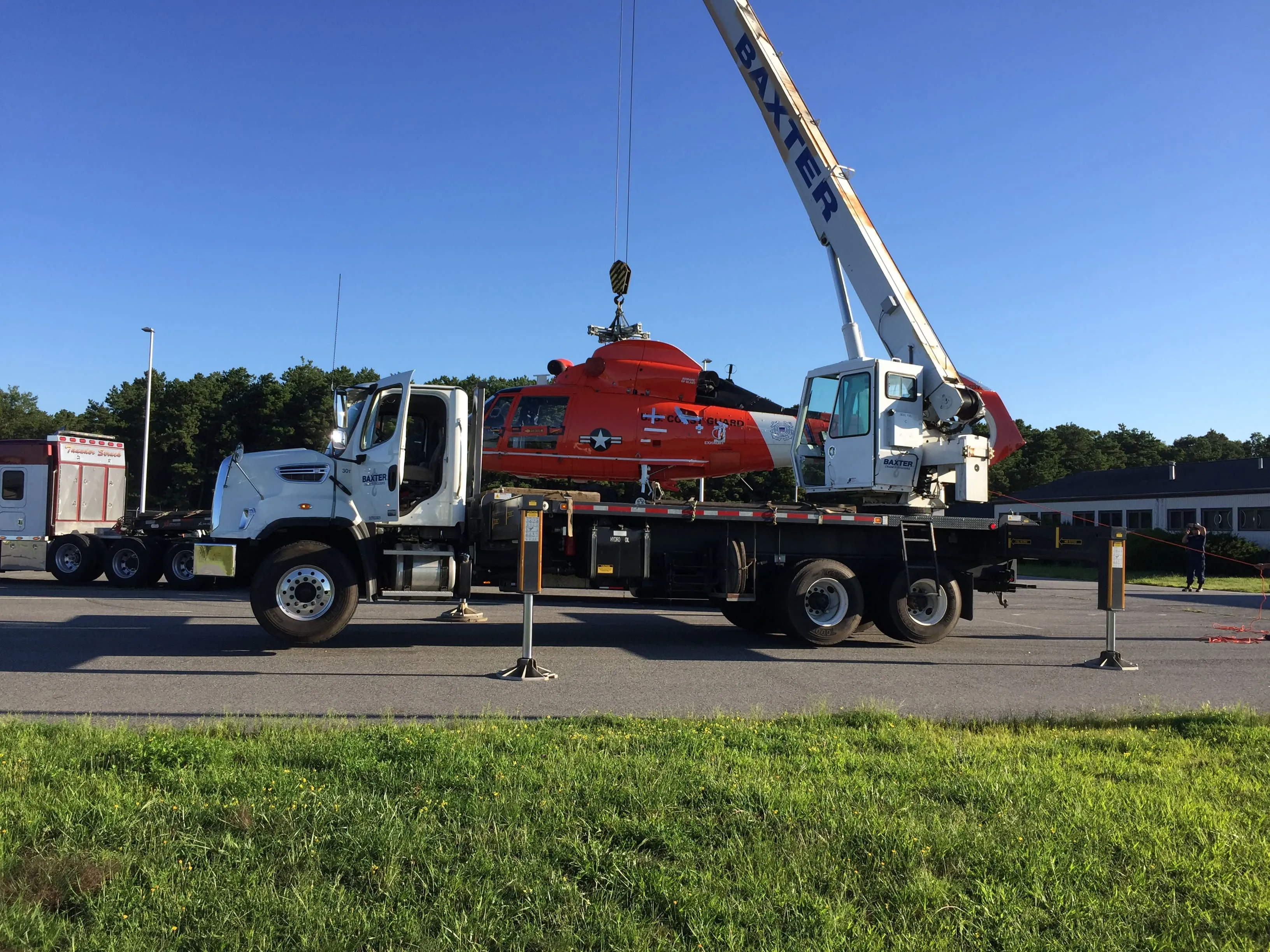 White crane truck labeled Baxter lifting a red and white Coast Guard helicopter on a paved surface with grass in the foreground.