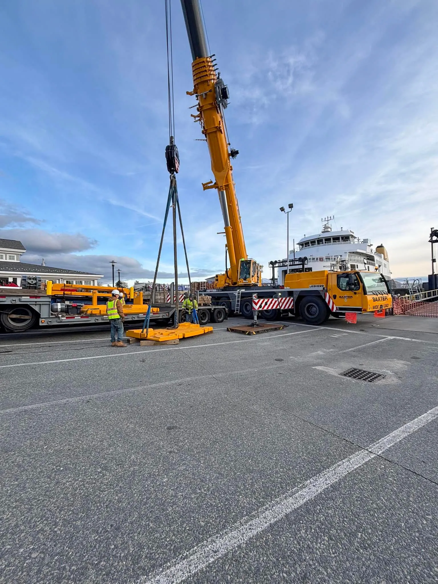 Yellow Baxter Liebherr TLM 1250 crane lifting equipment with workers guiding on a dock near a ferry.