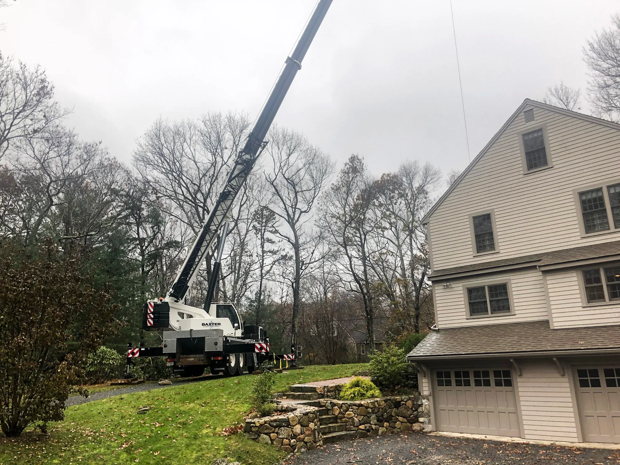 Mobile crane with extended boom parked on a grassy slope near a beige house with multiple windows and garages.