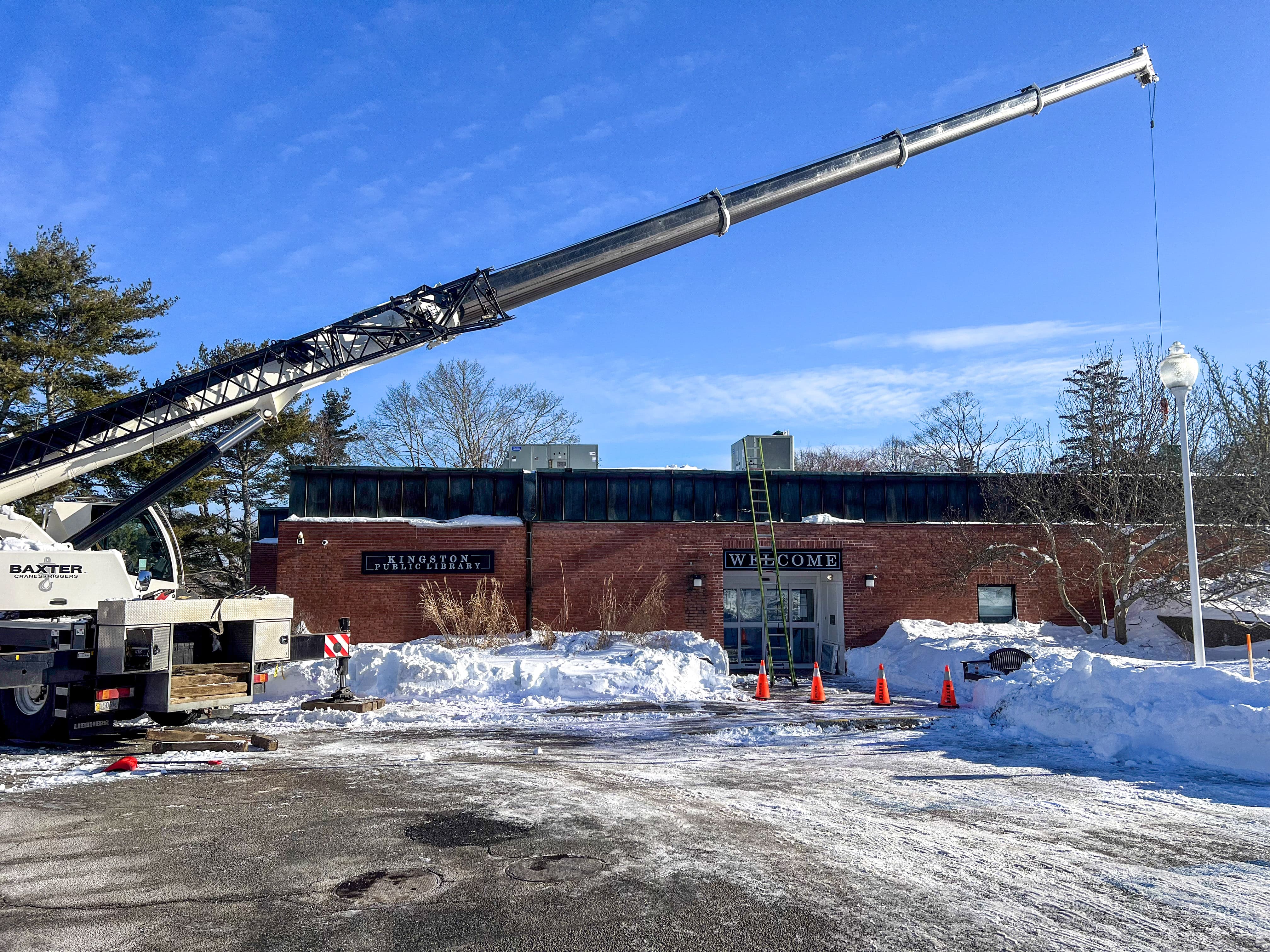 Large crane extended over the snow-covered Kingston Public Library building on a clear winter day.