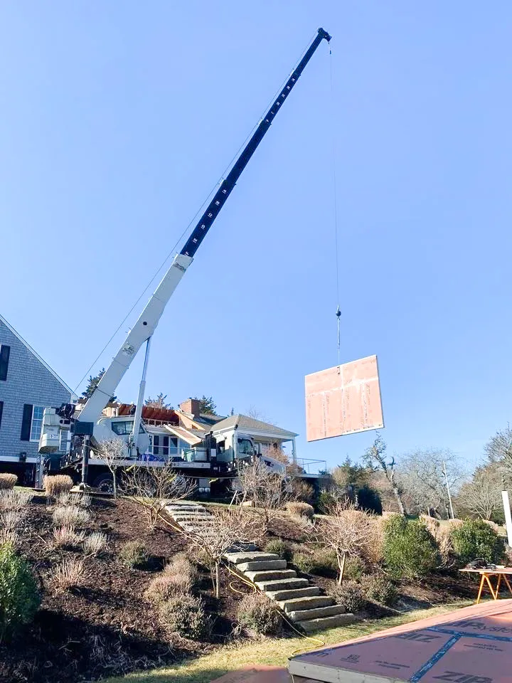 Large crane lifting a wooden panel above a residential construction site on a sunny day.