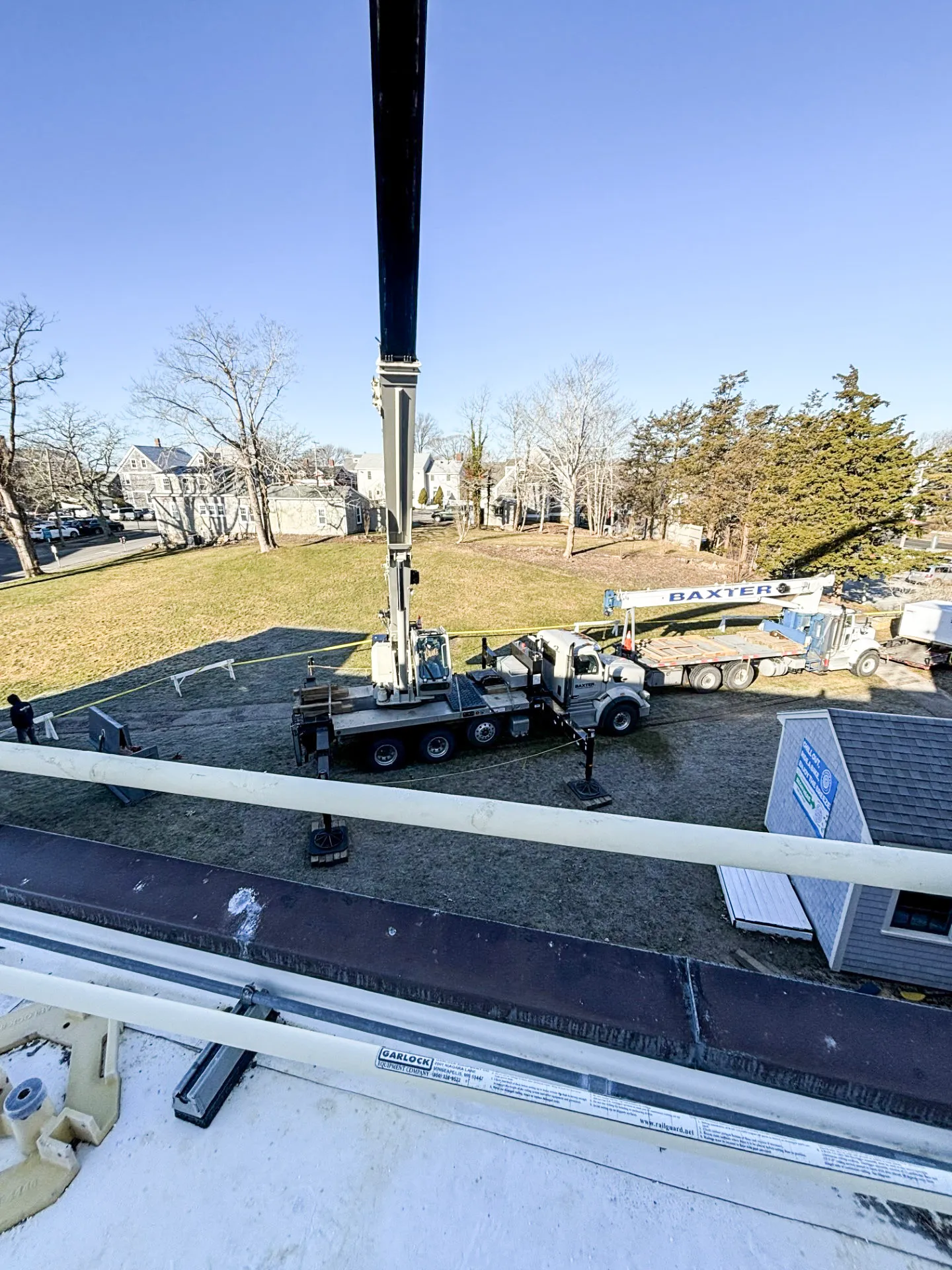 Overhead view of a large crane and a flatbed truck with 'Baxter' logo parked on a grassy area near houses and trees under a clear blue sky.