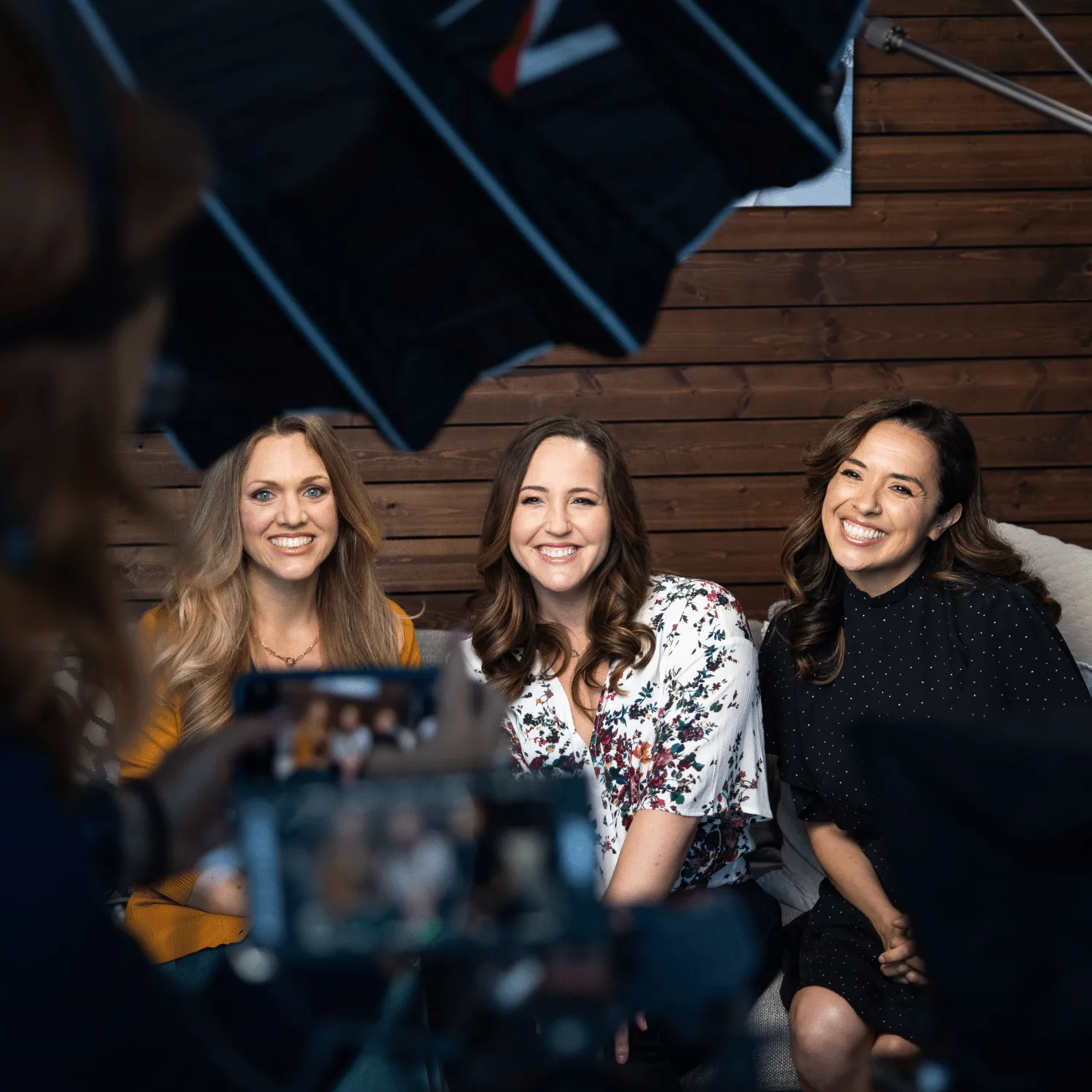 Three smiling women seated on a couch posing for a photo in a studio setting with studio lights and camera visible.