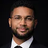 Man with short curly hair, beard, glasses, and wearing a suit and tie against a dark background.