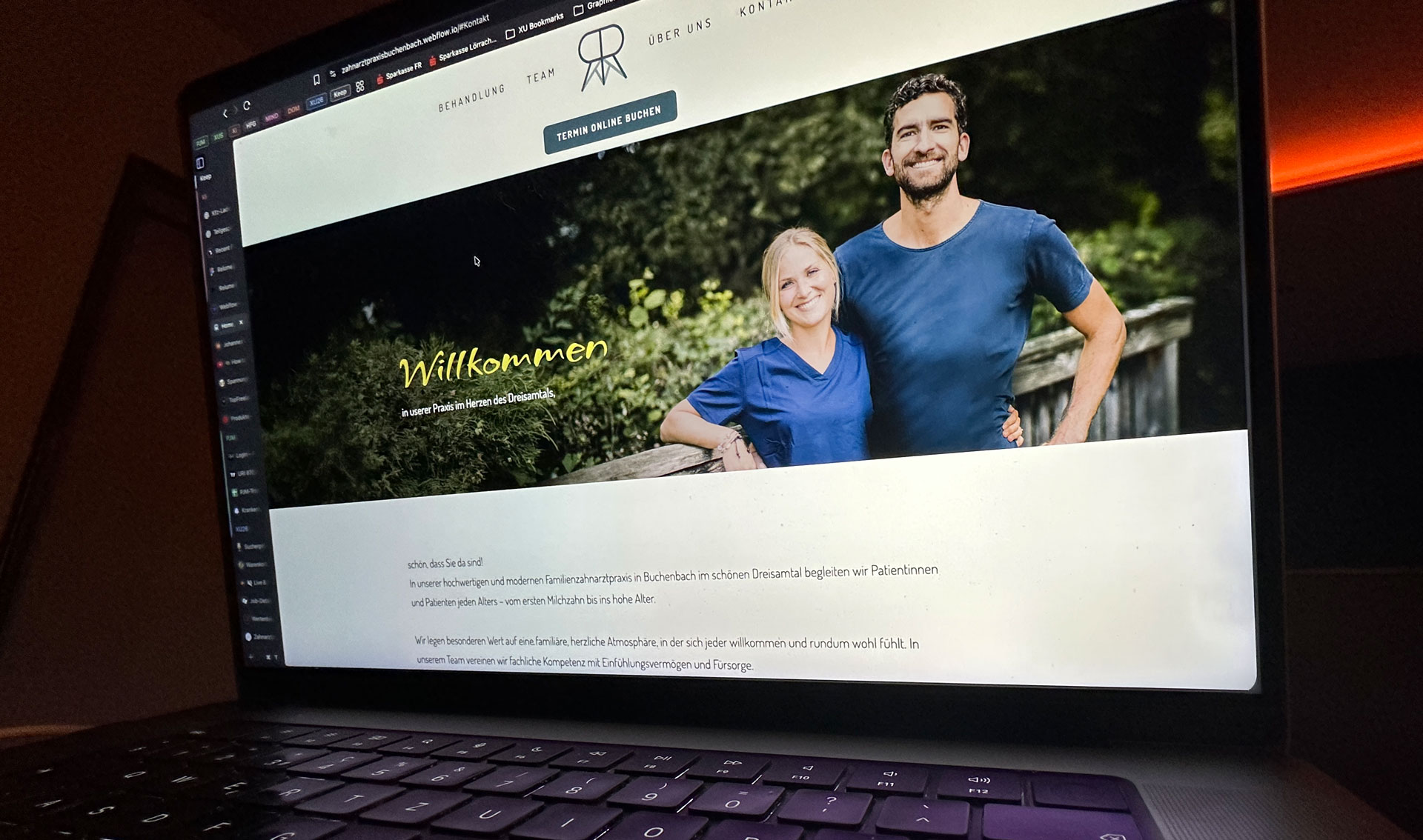 Laptop screen shows a dental practice website with a smiling man and woman in blue scrubs standing outdoors beside a wooden fence with greenery in the background.