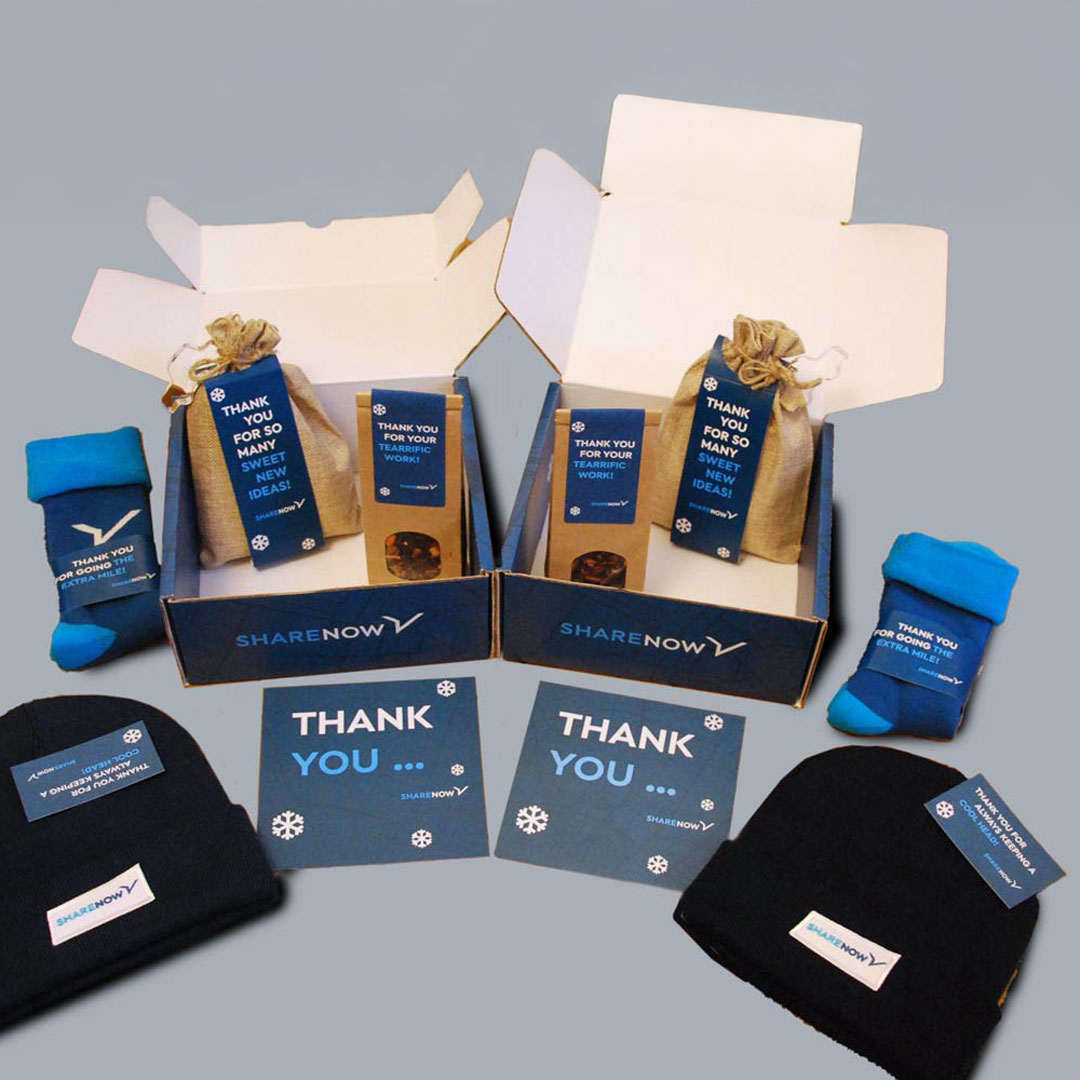 Zwei geöffnete Geschenkboxen mit SHARENOW Logo, jeweils mit blauen Dankeskarten, Socken, schwarzen Mützen und kleinen Geschenktüten.