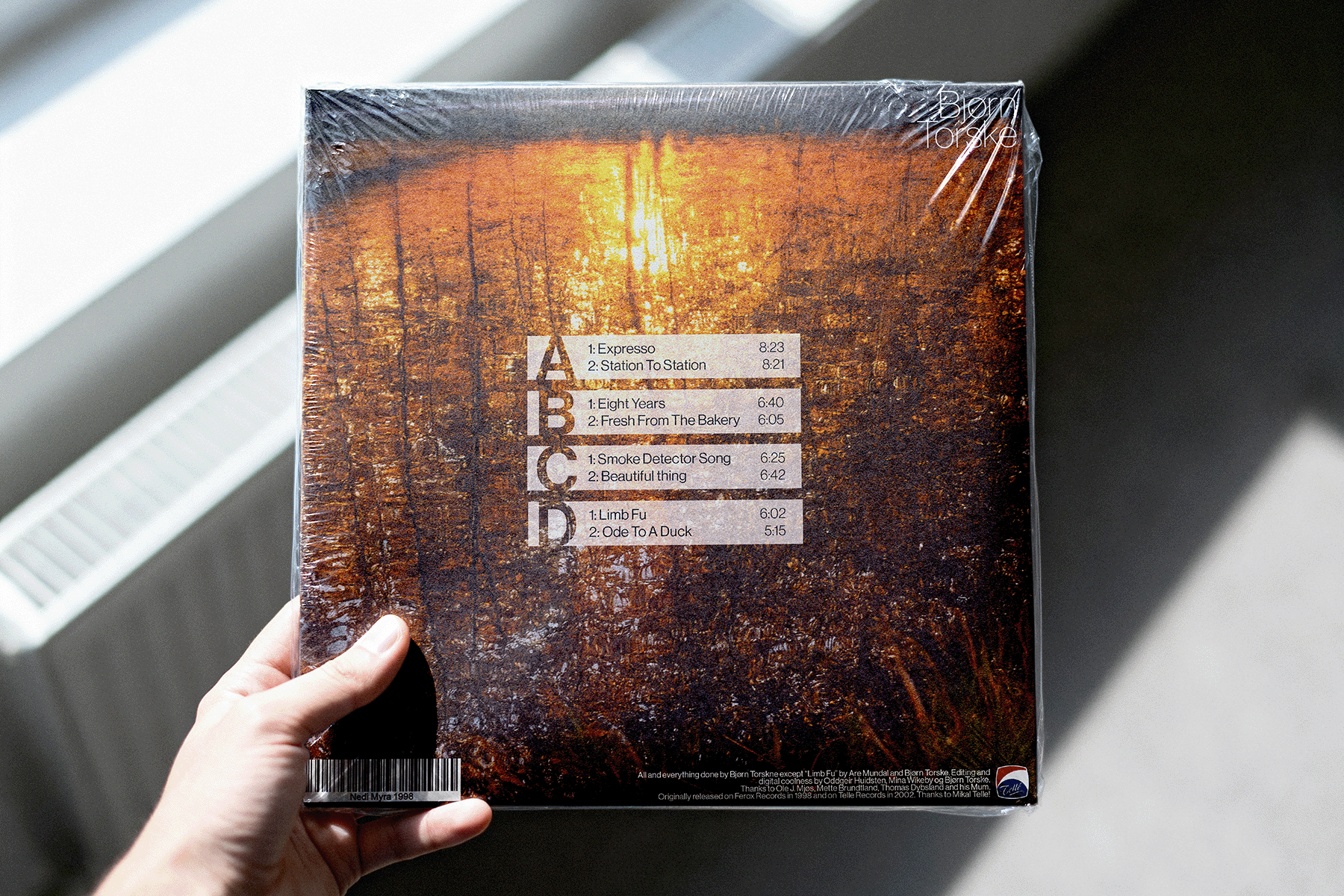 Hand holding a shrink-wrapped music album with tracklist and credits over a textured brown background.
